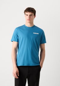T-shirt in cotone blu con maniche corte. Presenta un collo a giro e un logo bianco "Calvin Klein" sul lato sinistro del petto. Design semplice.