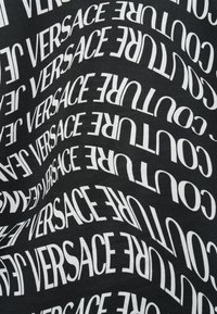 Čierna látka s bielym zakriveným opakujúcim sa textom "VERSACE JEANS COUTURE" v rôznych orientáciách.