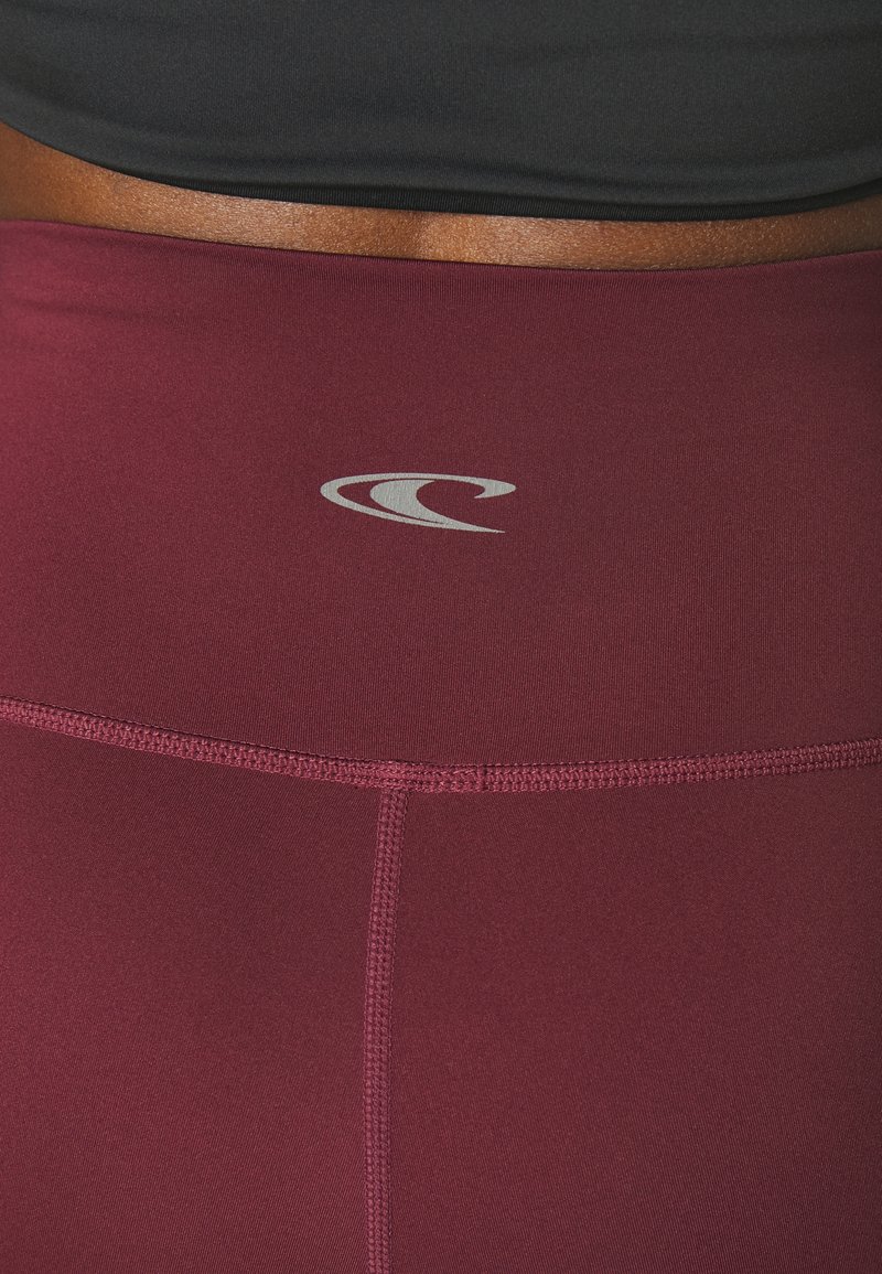 Bordeauxfarbene, hochgeschnittene Leggings mit sichtbaren Nähten und einem kleinen grauen Wellenlogo am Bund unter einem schwarzen Cropped Top.