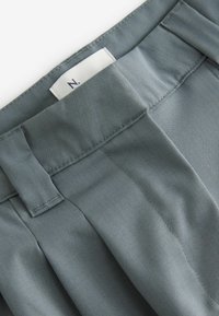 Pantaloni in tessuto grigio con pince, caratterizzati da una texture liscia e un'etichetta nella cintura. Il design include cuciture rifinite con cura.