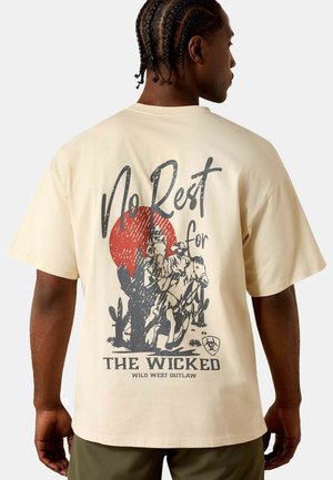 Man with braided hair wearing beige tee showing back design of a horseback rider, cactuses, red sun, and text "No Rest for The Wicked Wild West Outlaw."