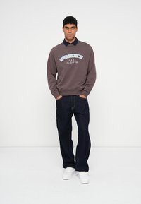 Hnedá mikina s logom "Tommy Jeans" v bielej a svetlomodrej farbe, skombinovaná s tmavými džínsami a bielymi teniskami. Model stojí na jednoduchom pozadí.