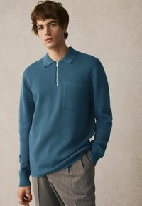 Teal langærmet poloshirt med lynlås krave, tekstureret stof og enkelt brystlomme, parret med grå stribede bukser.
