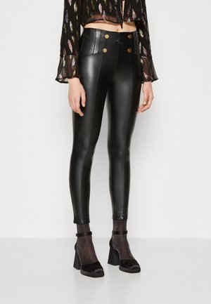 Kvinde iført sorte, højtaljede leggings af kunstlæder med guldknapper, gennemsigtig mønstret bluse og glitrende sokker med sorte blokhæle.