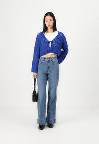 Cropped blue crochet cardigan with a tie closure, paired with high-waisted, flared denim jeans. The model holds a black handbag.