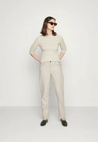 Suéter ajustado de color beige claro, pantalones crema ajustados y zuecos de color verde oscuro. La modelo está de pie con las manos en las caderas, usando gafas de sol oscuras.