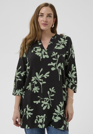 Kobieta o długich brązowych włosach, ubrana w czarną tunikę z zielonymi liściastymi wzorami i jasnoniebieskie dżinsy, stojąca na jednolitym tle.