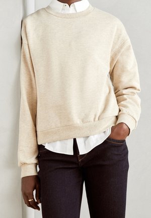 Persoon draagt een crèmekleurige sweater met ronde hals over een wit overhemd, gecombineerd met donkerblauwe spijkerbroek, met één hand in de zak en leunend tegen een muur.