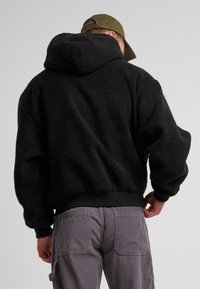 Haut à capuche en polaire noire avec un extérieur texturé, associé à un pantalon en velours côtelé gris. Les poignets et l'ourlet sont bordés d'une bande lisse.