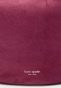 Sac en daim bordeaux avec une texture lisse, affichant le nom de la marque "kate spade NEW YORK" en blanc. Design minimaliste sans accents supplémentaires.