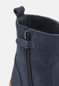 Bota de tornozelo em camurça azul-marinho com fecho de zíper, superfície texturizada e um puxador no colarinho. Apresenta sola castanha em contraste.