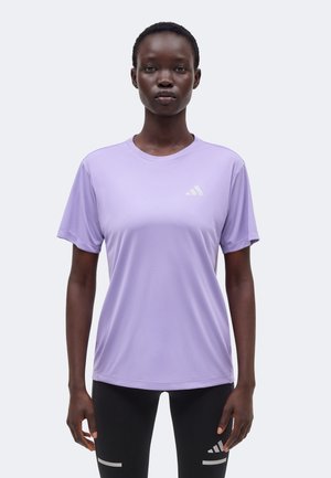 Nainen seisoo kasvot eteenpäin, yllään vaalean violetti Adidasin lyhythihainen paita ja mustat leggingsit, joissa heijastavia yksityiskohtia.