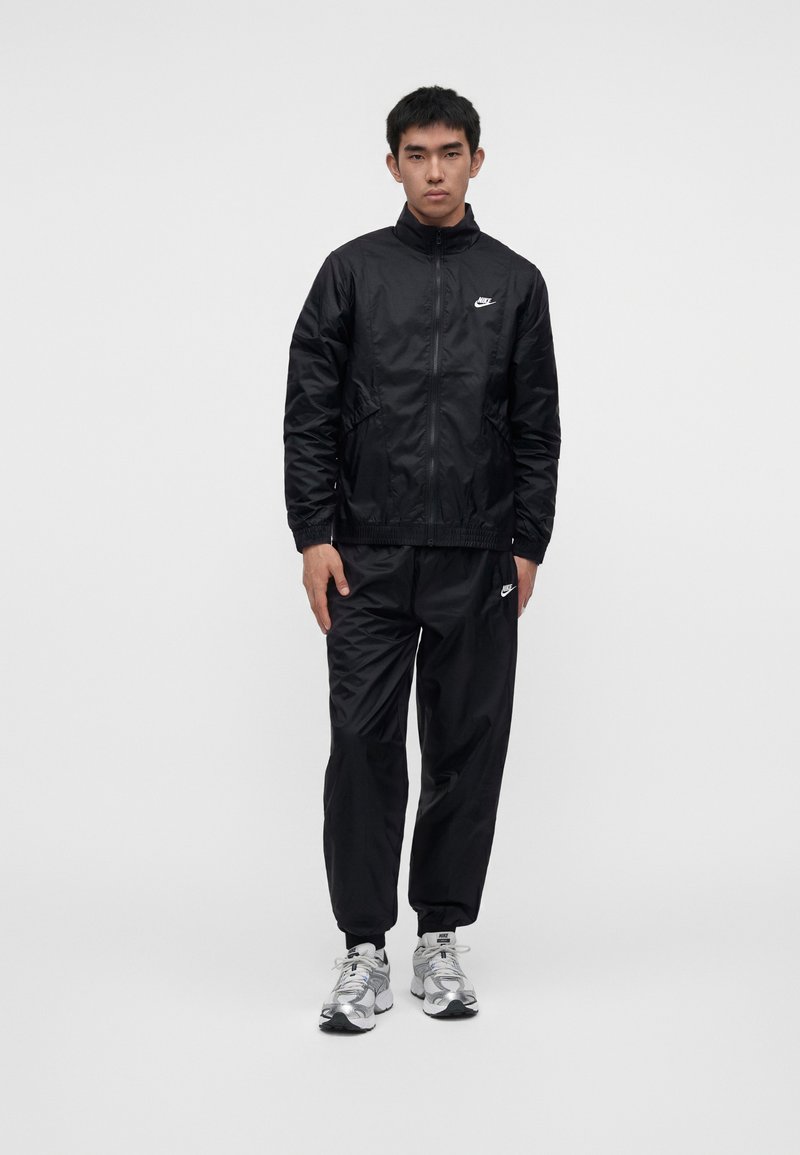 Νέος άνδρας που φοράει μαύρο φόρμα της Nike με λευκά λογότυπα Nike και ασημένια-λευκά αθλητικά παπούτσια, στέκεται μπροστά σε απλό ανοιχτό φόντο.