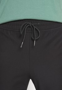 Gros plan sur un pantalon de survêtement noir avec une taille à cordon noué, porté avec une chemise verte.