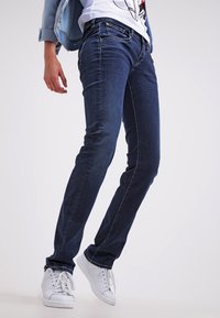 Jeans ajustados de denim azul oscuro, con cintura de altura media, cinco bolsillos y costuras en contraste sutil. Combinados con zapatillas blancas.