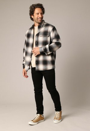 Chemise en flanelle à carreaux noir et blanc avec fermeture à boutons, associée à un t-shirt beige, un jean skinny noir et des baskets montantes beige.