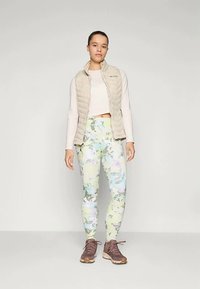 Lagani bež kvasasti prsluk preko bijelog kratkog majice, uparen sa višebojnim pastelnim tajicama i smešnim sportskim cipelama.