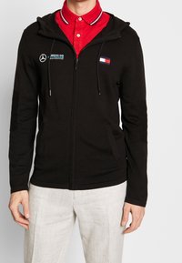 Uomo con felpa nera con zip con i loghi Mercedes AMG Petronas Motorsport e Tommy Hilfiger, sopra una camicia rossa con colletto, e pantaloni grigio chiaro.