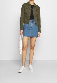 Olivgrüne Jeansjacke, schwarzes T-Shirt, blauer Jeansrock mit ausgefranstem Saum, weiße Handtasche und weiße Sneakers mit strukturierten Sohlen.