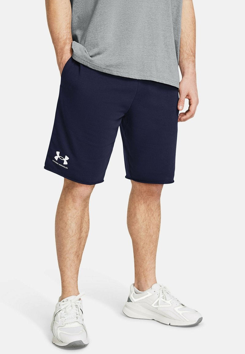 Marina blå sportshorts med elastisk midjeband, sidofickor och vitt Under Armour-logotyp på fållen. Bärs tillsammans med en grå t-shirt och vita sneakers.