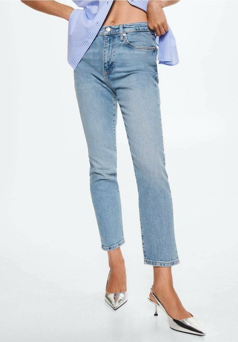 Femme portant un jean droit taille haute bleu clair, chemise bleue partiellement rentrée et chaussures à talons hauts argentées pointues, debout.