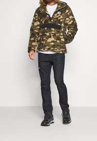 Man med grön och beige kamouflage fleece-tröja, svarta byxor och svarta vandringsskor som står mot en vit bakgrund.