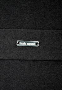 Czarna tkanina o widocznej teksturze; na niej srebrna metka z napisem "kasia zapata," przyczepiona za pomocą prostokątnego otworu.