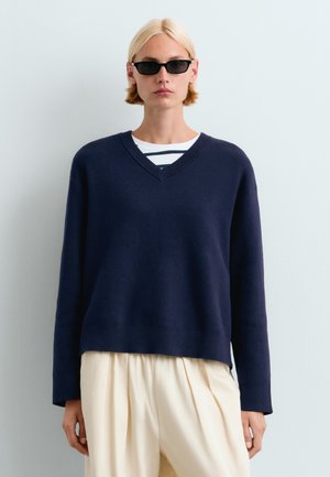 Personne blonde portant des lunettes de soleil rectangulaires noires, un pull bleu marine à col en V sur une chemise rayée, et un pantalon ample couleur crème sur un fond uni.