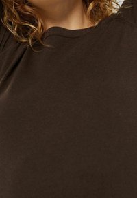 Osoba z kręconymi brązowymi włosami, ubrana w ciemno-brązowy sweter typu crew neck, przycięta od szyi do górnej części torsu na jednolitym tle.