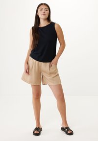 Kvinna som står med en hand i fickan, iklädd en ärmlös svart topp, beige shorts, svarta slidesandaler och ett guldfärgat halsband mot en vit bakgrund.