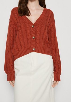 Frau trägt eine rostfarbene, kurz geschnittene Strickjacke mit Zopfmuster und Schildpattknöpfen über einem hoch taillierten weißen Rock, stehend vor einem einfarbigen Hintergrund.