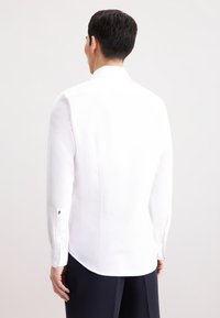 Camicia bianca a maniche lunghe con design a colletto, texture liscia, aperture laterali e polsini con bottoni, abbinata a pantaloni scuri.