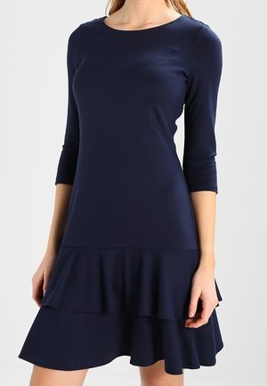 Femme portant une robe bleu marine à manches longues avec une jupe à volants superposés, debout devant un fond uni clair.