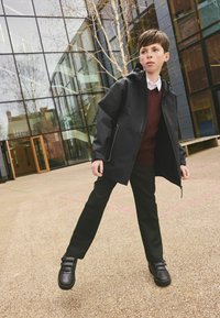 Garçon portant une veste noire, un pull bordeaux, un pantalon noir et des chaussures noires, debout les jambes écartées près d’un bâtiment moderne en verre.