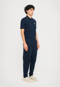Marineblå polo shirt med guld accenter og logo, parret med matchende marineblå joggers. Modellen har kremfarvede sneakers på og står foran en ensfarvet baggrund.