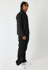Giacca nera con colletto alto e chiusura a zip, abbinata a pantaloni cargo coordinati con tasche e bottoni a pressione. Scarpe da ginnastica bianche completano l'outfit.