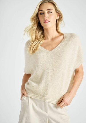 Vrouw met lang blond haar, gekleed in een beige, zachte trui met V-hals en crème wijde broek, poserend tegen een eenvoudige lichte achtergrond.