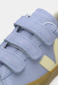 Gros plan sur une chaussure de sport en daim bleu clair avec trois bandes velcro et une semelle en caoutchouc beige.