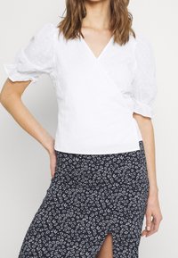 Haut blanc drapé avec manches bouffantes et pois texturés, associé à une jupe bleu marine à imprimé floral avec une fente haute sur la cuisse.