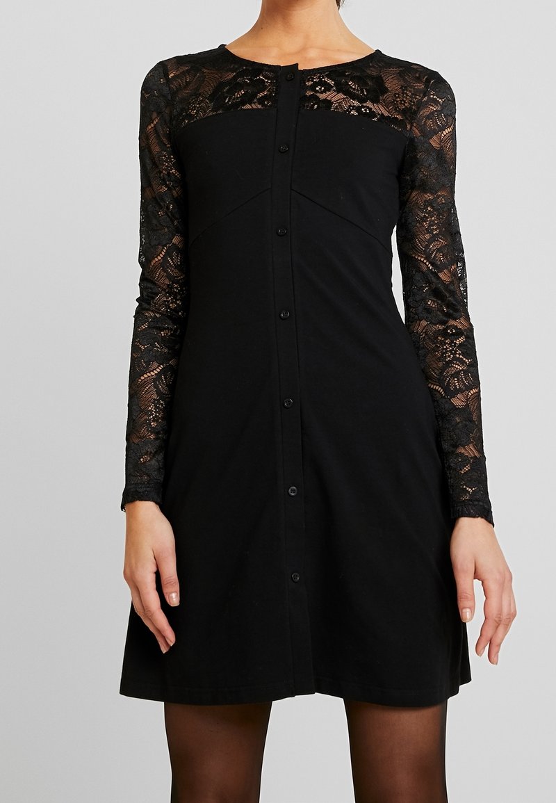 Femme portant une robe noire avec des manches longues en dentelle et un haut en dentelle sur la poitrine, avec des boutons devant et un col rond.