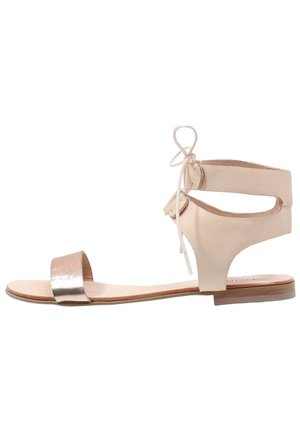Beżowe skórzane sandały z metaliczną srebrną przednią taśmą i wiązaniami wokół kostki, o otwartym fasonie i płaskiej drewnianej podeszwie.