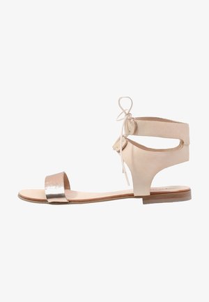 Sandale en cuir beige avec une sangle avant argentée métallique et des liens à la cheville, au design ouvert et à semelle en bois plate.