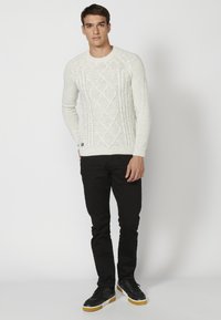 Cremfarbener Strickpullover mit Diamantmustern, gerippten Bündchen und Saum, kombiniert mit schwarzen Slim-Fit-Hosen und schwarzen Sneakers.