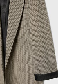 Blazer i tan- och svart hundtandsmönster med urklippt slag, en ficka och svarta satinklemmar på ärmarna. Texturerat tyg.