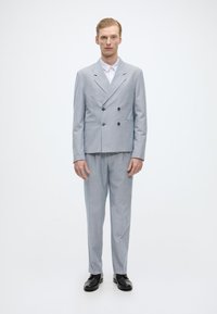 Ανοιχτό γκρι σακάκι κοστουμιού με διπλό κούμπωμα και καπάκια, συνδυασμένο με αντίστοιχες παντελόνες. Συμπληρώματα: λευκό πουκάμισο, μαύρα παπούτσια, κομψή εφαρμογή.