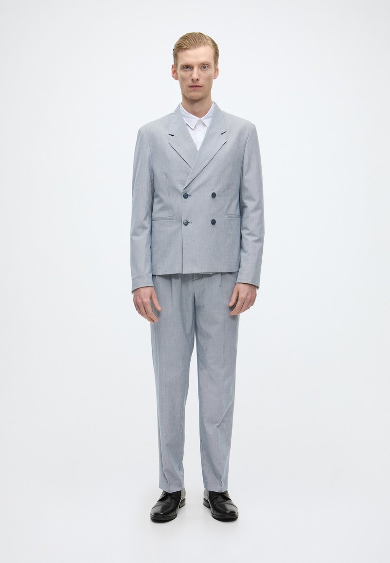 Ανοιχτό γκρι σακάκι κοστουμιού με διπλό κούμπωμα και καπάκια, συνδυασμένο με αντίστοιχες παντελόνες. Συμπληρώματα: λευκό πουκάμισο, μαύρα παπούτσια, κομψή εφαρμογή.