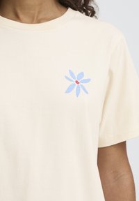 Kremna bombažna majica z modrim cvetličnim vzorcem s rdečim sredinskim delom, okroglim izrezom in kratkimi rokavi. Mehka tekstura z udobnim krojem.