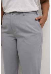 Pantaloni grigio chiaro con una texture liscia, dotati di chiusura con bottoni, passanti per cintura e tasche laterali. Vestibilità sartoriale e dettagli minimalisti.