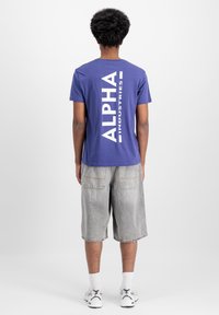 Lila crew-neck póló fehér "ALPHA INDUSTRIES" felirattal a hátsó részen, világosszürke farmer rövidnadrággal és fehér sportcipővel párosítva.