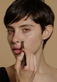 Un pinceau de maquillage est utilisé sur le visage d'un modèle, avec des cheveux courts et foncés et une peau lisse, appliquant un produit de couleur claire sur le nez.
